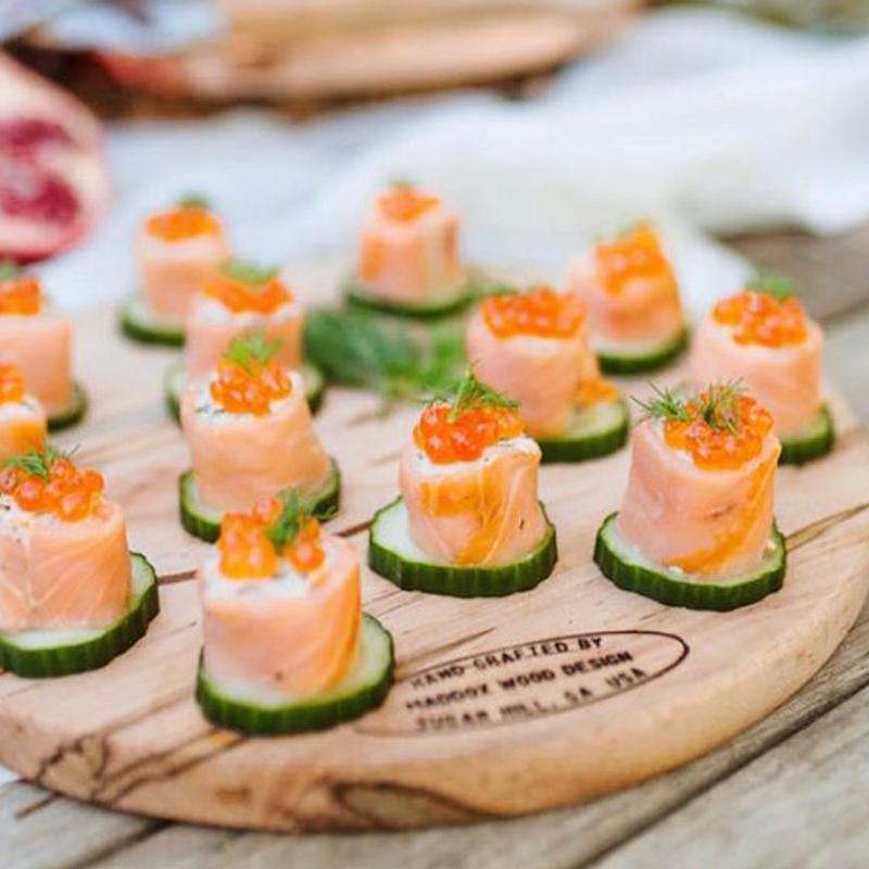 Smoked salmon rolls topped with salmon roe and dill, each set on a cucumber round across a round wooden board.