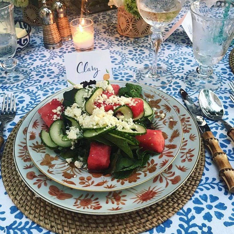 A watermelon, cucumber and feta salad plated on patterned china at a laid table, with a handwritten place card behind it.