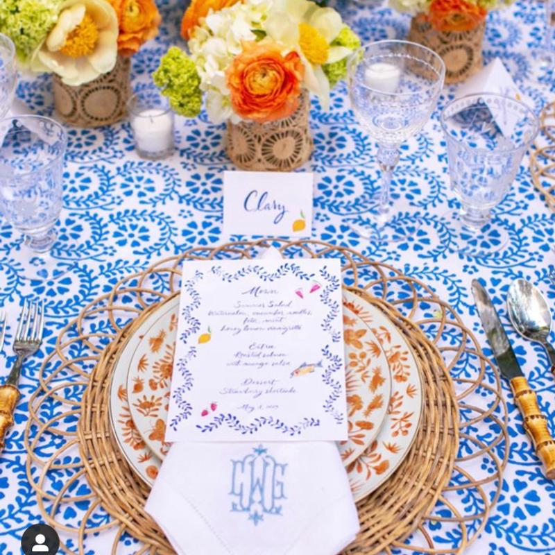 A wedding place setting on blue block-print linen: a handwritten Minston’s menu card on patterned china over a rattan charger, with a monogrammed napkin and cut glassware.